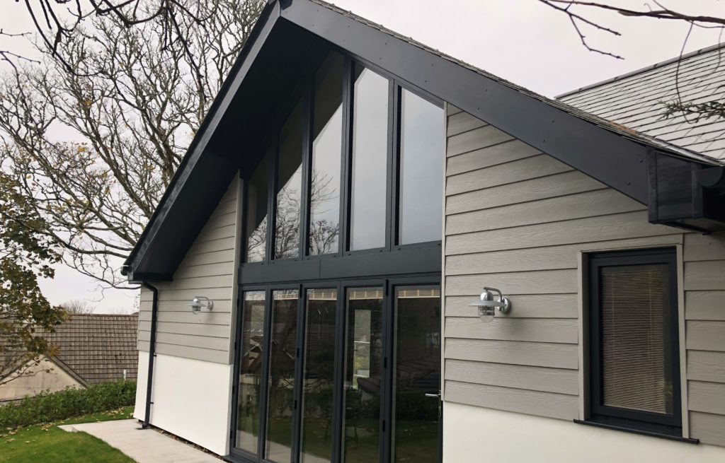 Modern house with large glass windows and a pitched roof. The exterior is grey, with surrounding trees indicating a serene setting.