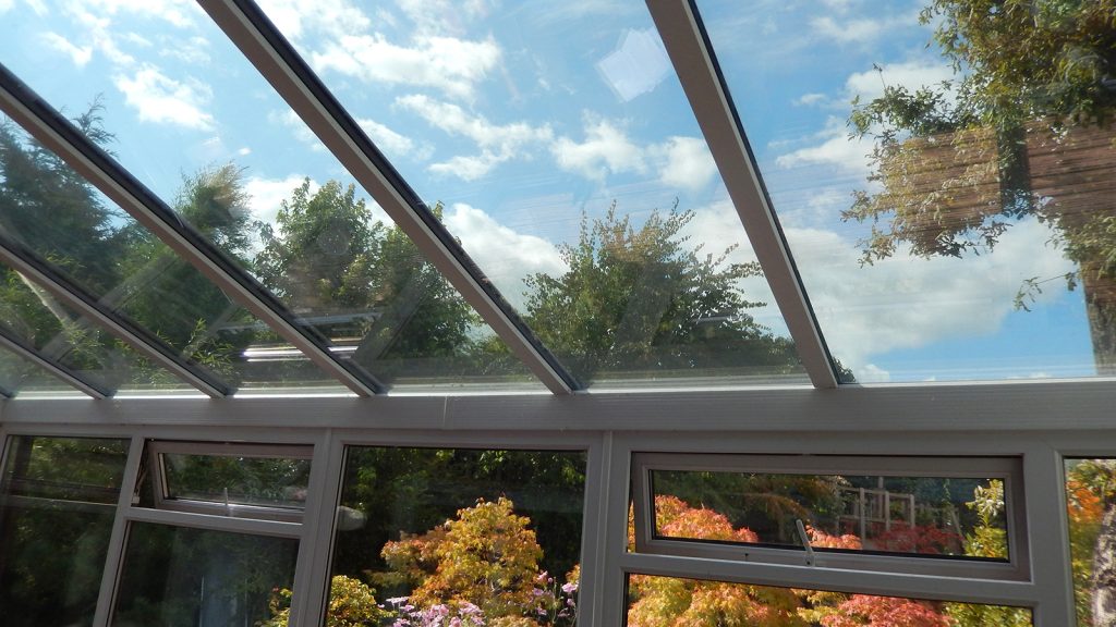 View from inside a conservatory with a glass ceiling, revealing green trees and colorful foliage below a bright sky and fluffy clouds.