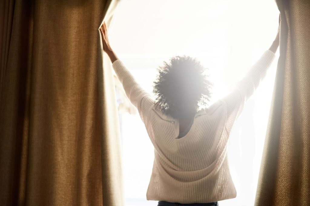 A person stands in front of a window, pulling apart thick curtains to reveal bright, natural sunlight.