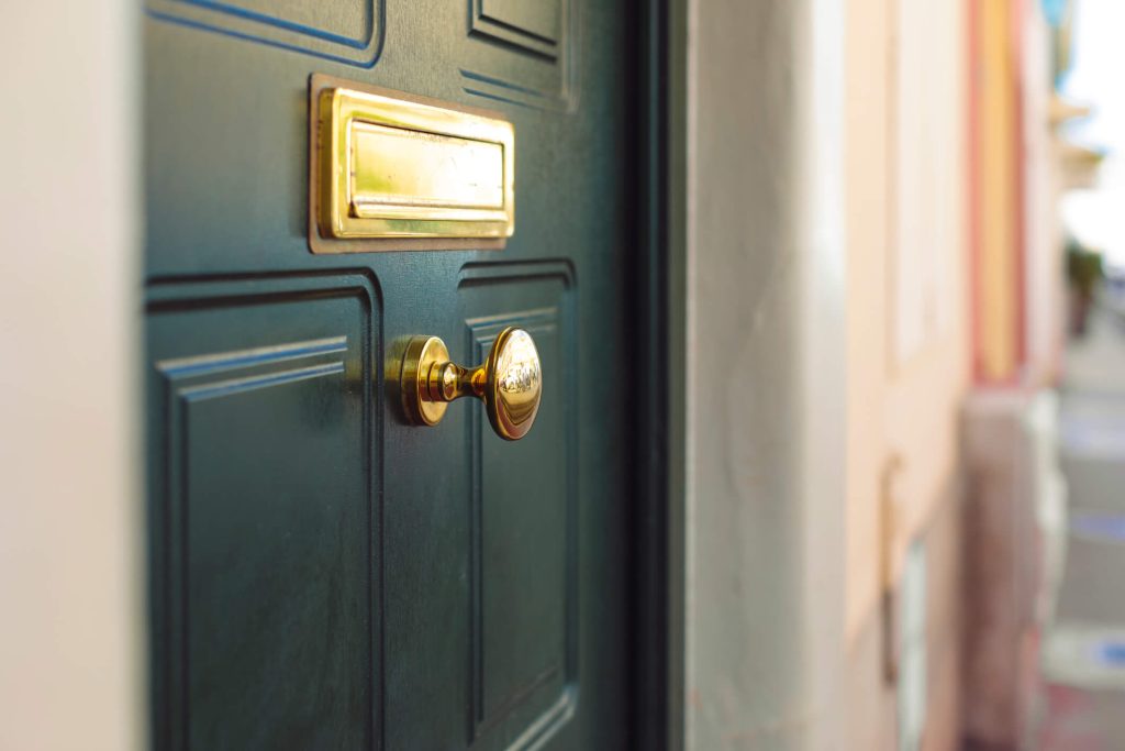 Close-up of a green door with a brass doorknob and mail slot, with a blurred pastel-colored building background.