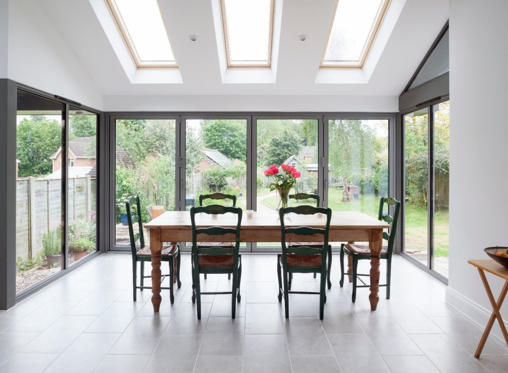 Dining room with a wooden table and six chairs, under skylights, next to glass doors that reveal a green garden outside.