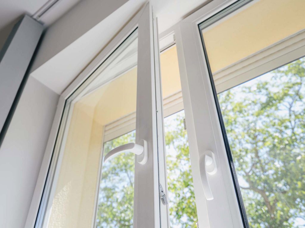 Open white-framed window with clear glass, revealing a sunny view of lush green trees.